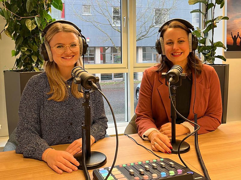 Zwei junge Frauen sitzen mit Kopfhörern vor Mikrofonen und lächeln in die Kamera